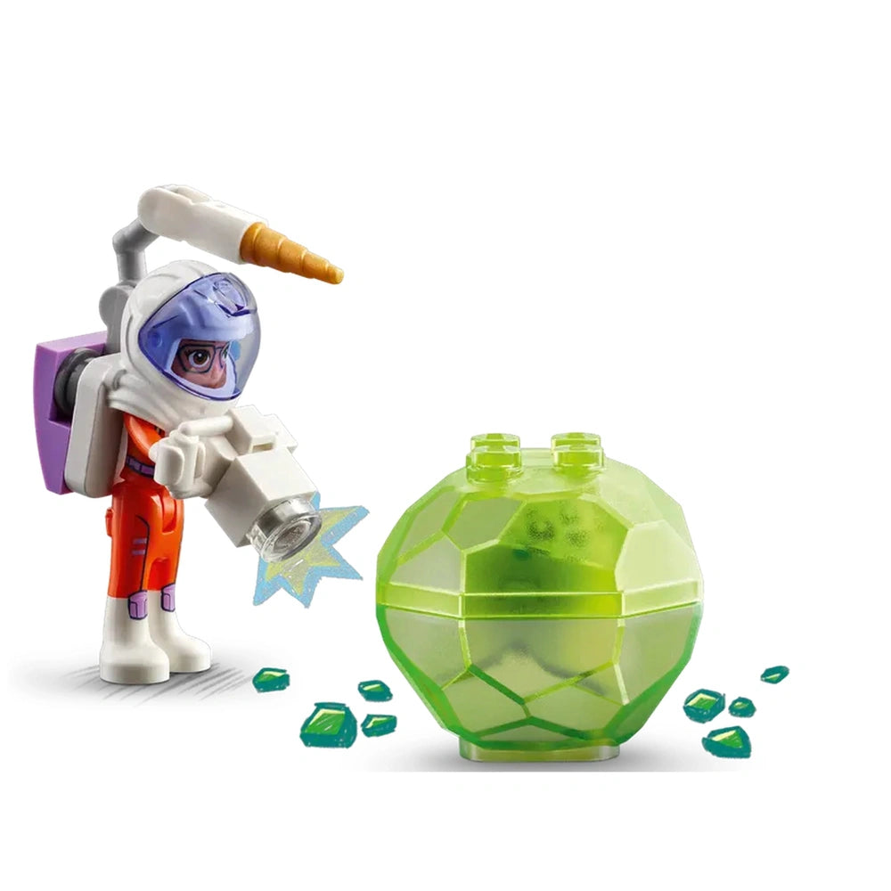 Astronaut mini-doll in a bright orange suit, holding a device with a star effect, stands next to a green, gem-like structure with scattered translucent pieces.