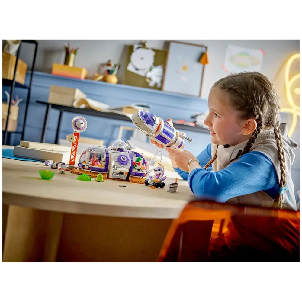 A child with braided hair joyfully plays with the LEGO® Friends Mars Space Base set, holding a rocket in one hand while exploring the colorful base and mini-dolls on a round table.