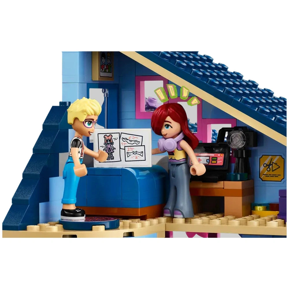 Two LEGO mini-dolls, one blonde and the other with red hair, interact inside a colorful house. The boy gestures towards a drawing on a table, while the girl looks on, wearing headphones. The room features bright wall decorations, a small lamp, and additional accessories, creating an inviting atmosphere for imaginative play.