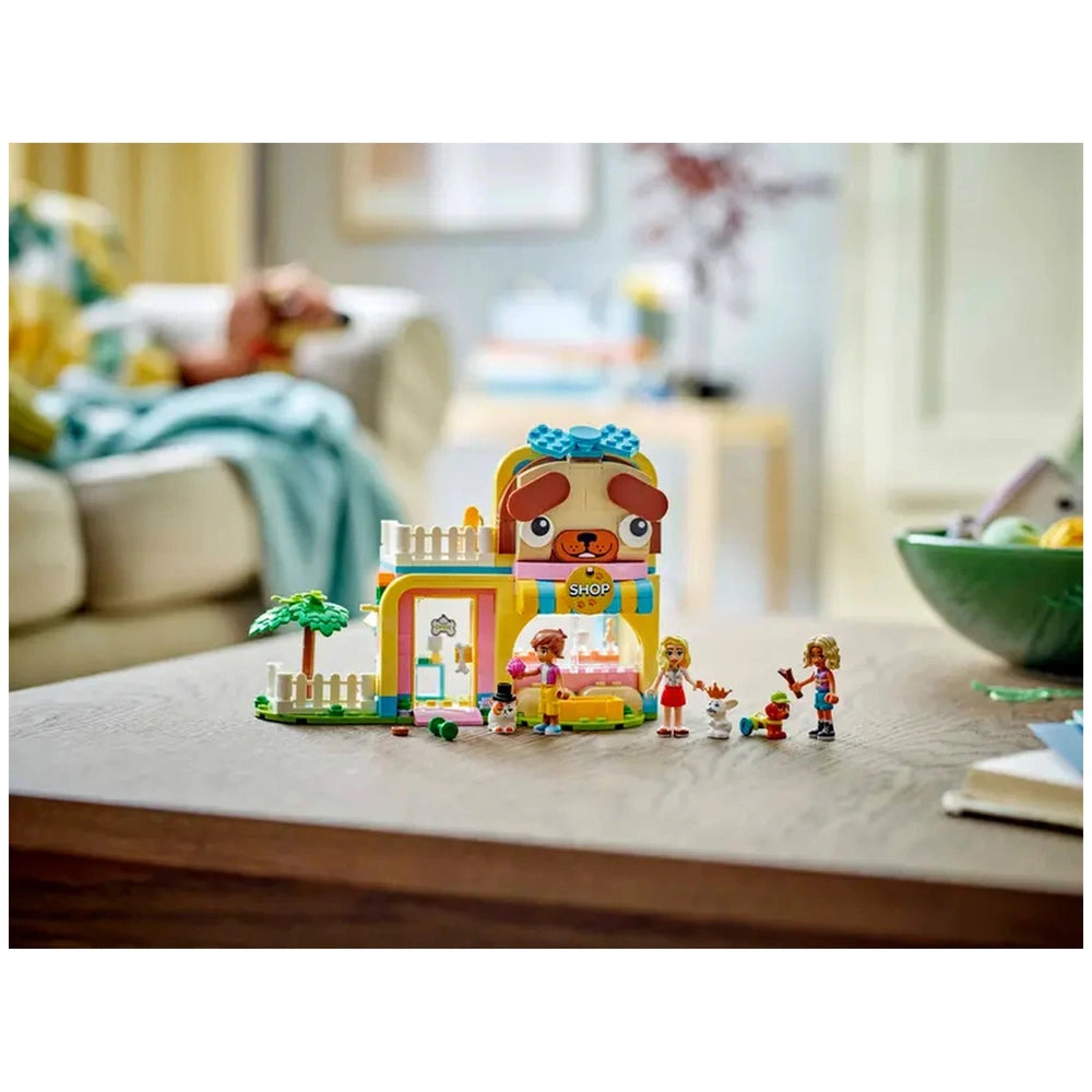 Colorful LEGO® Friends Pet Accessories Shop set featuring three minidolls and animal figures. The shop has a playful dog face and includes grooming accessories, surrounded by a tree and fence on a wooden table.