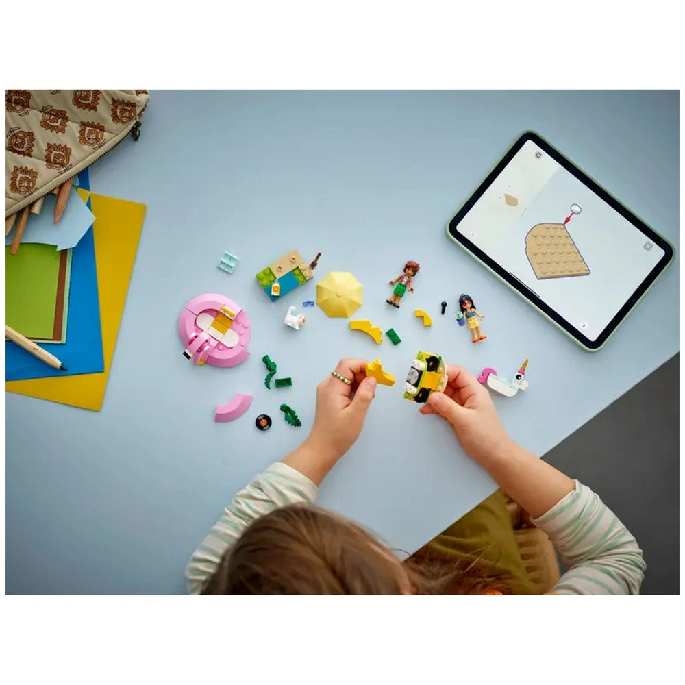 Child's hands assemble a yellow LEGO car amidst colorful pieces, a pink floatie, and two minidolls, with a tablet displaying building instructions nearby.