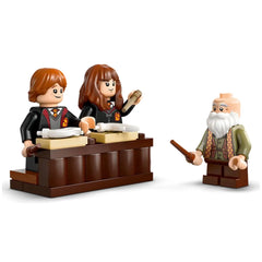 Two LEGO figures, Ron and Hermione, are seated at a brown desk with scrolls, while Professor Flitwick stands nearby, holding a wand, in a classroom setup.