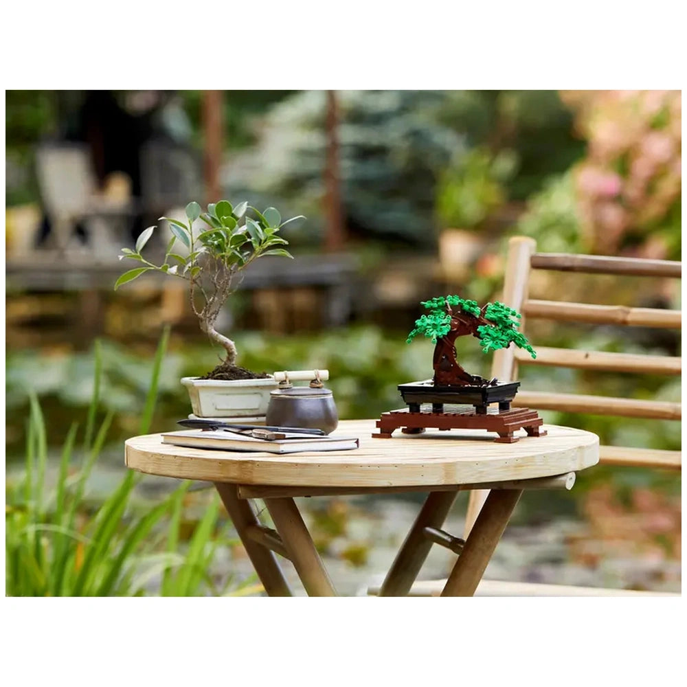Lego Bonsai Tree model set on a wooden table beside a real bonsai plant, with green leaves and a serene garden backdrop.