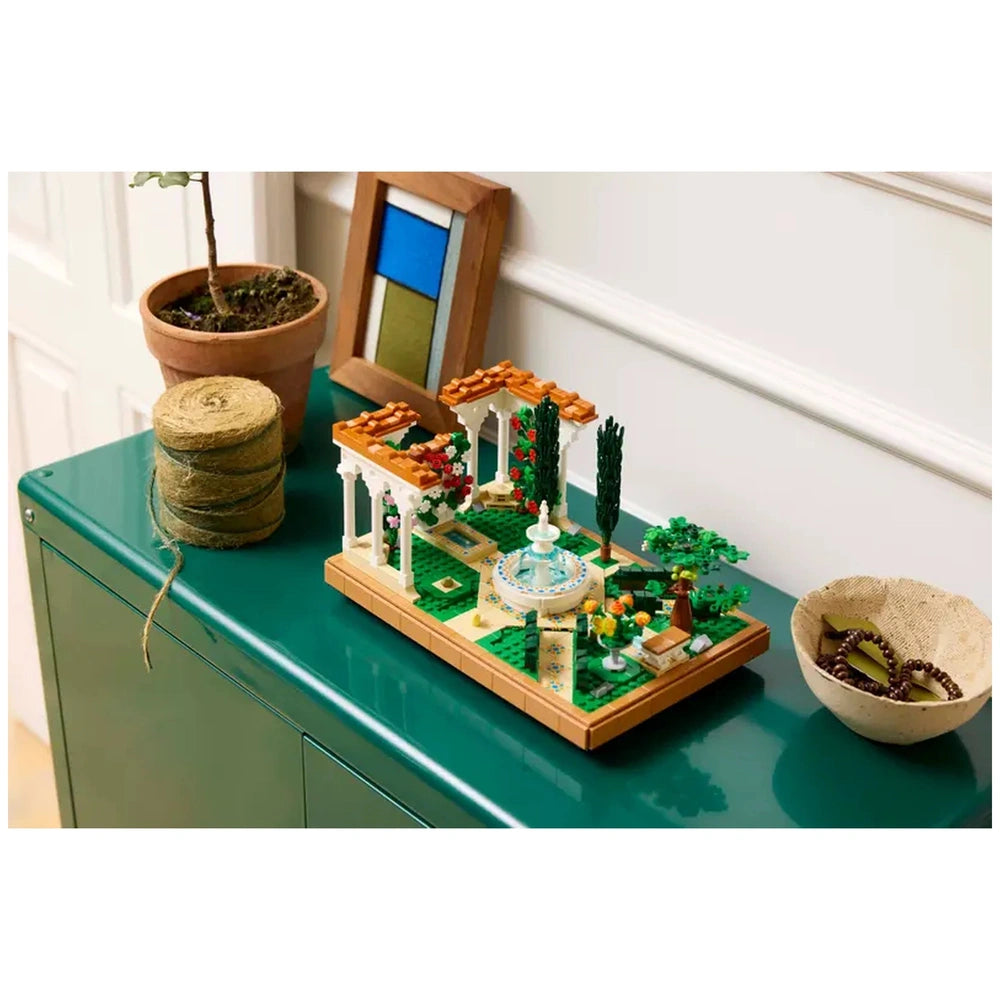 A detailed LEGO® Icons Fountain Garden construction set features a tiered fountain surrounded by colorful flowers, green trees, and decorative pathways. It is displayed on a green table next to a potted plant, a spool of twine, and a bowl with decorative items.
