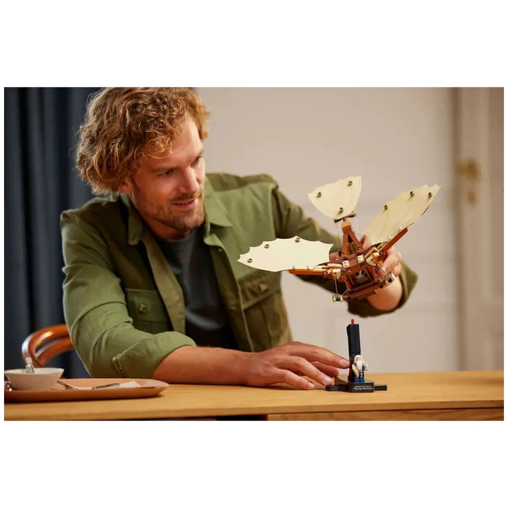 Adult male with curly hair smiles while assembling LEGO® Icons Leonardo da Vinci's Flying Machine on a table, showcasing intricate flappable wings.