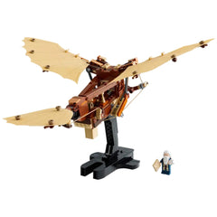 LEGO® Icons Leonardo da Vinci's Flying Machine model with tan flappable wings, mounted on a black stand, alongside a minifigure of da Vinci holding a design sketch.
