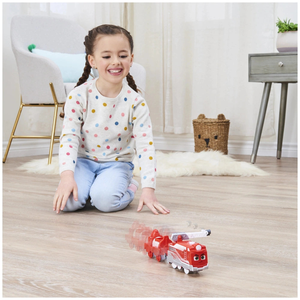 A young girl with braids smiles while sitting on a rug, playing with a colorful Mighty Express motorized train.