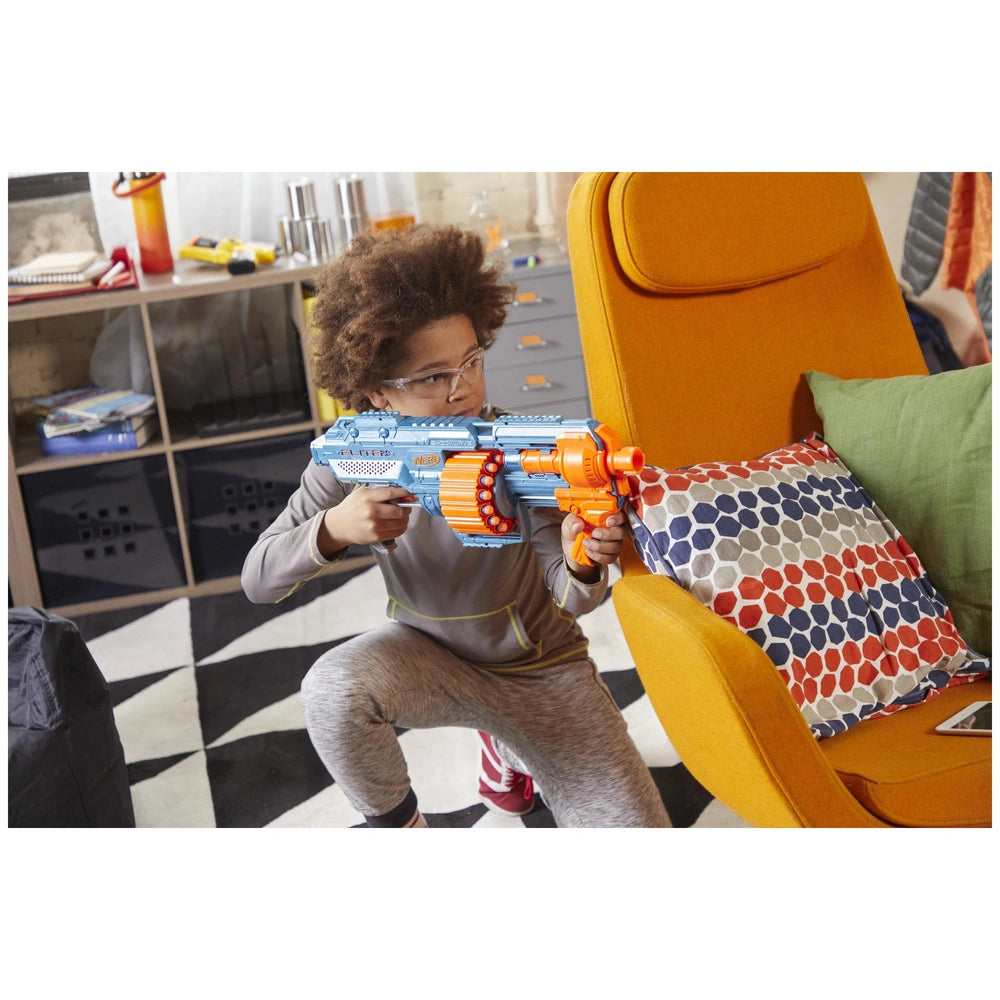 Child in gray shirt holds blue and orange Nerf Elite 2.0 Shockwave RD-15 Blaster while sitting on an orange chair with a pill