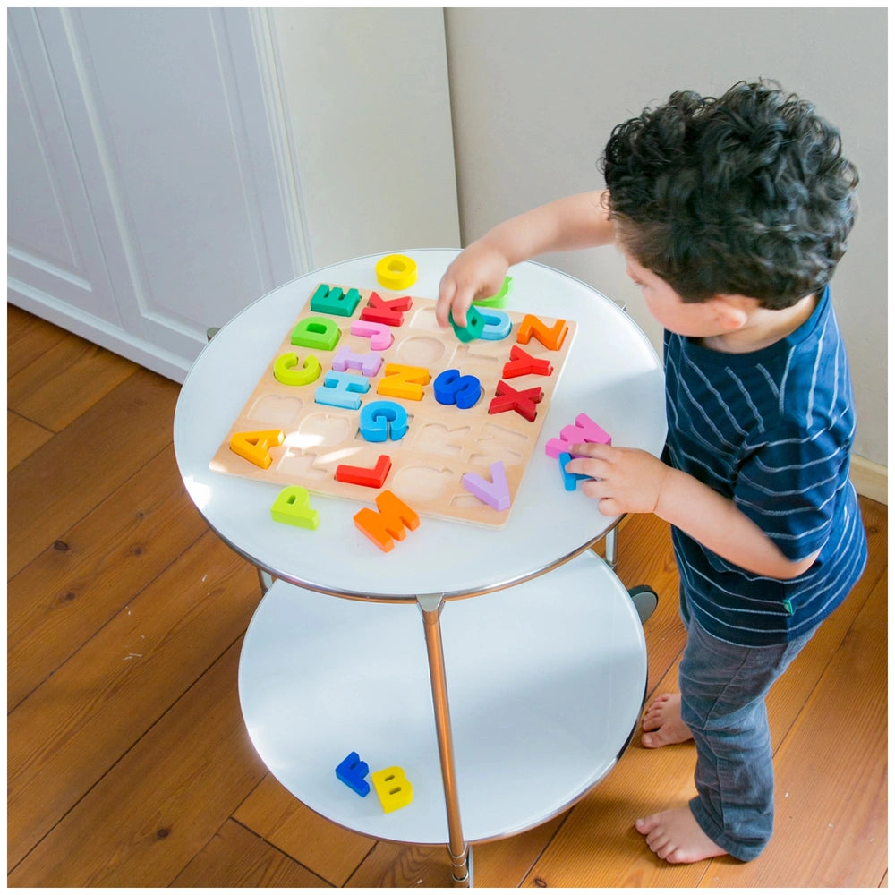 Uppercase Alphabet Puzzle by New Classic Toys, colorful letters on a round white table, child in blue shirt engaged in play.