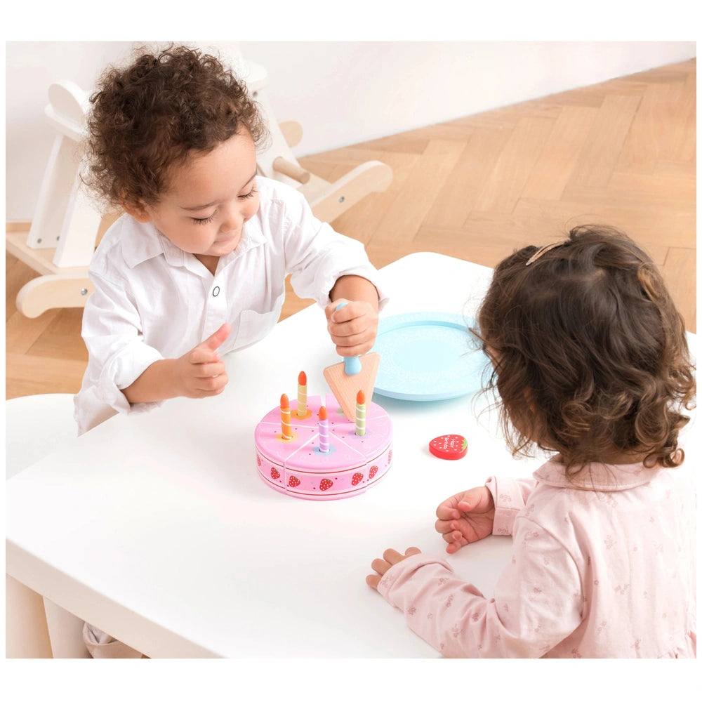 New Classic Toys Cutting Cake set on a wooden table with children playing, featuring colorful plastic cake and decorations.