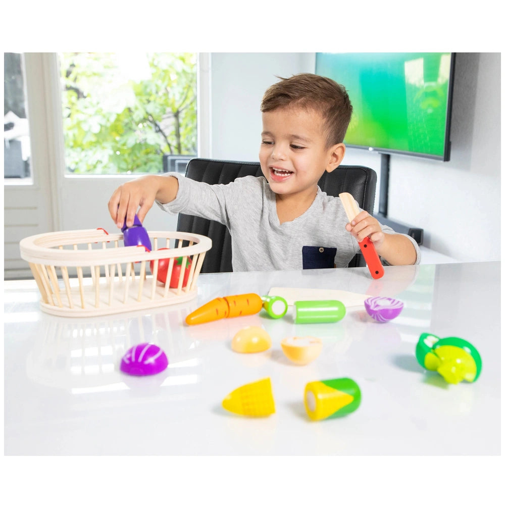 Young child in gray t-shirt seated at a white table with a wicker basket containing red and purple objects, holding green and