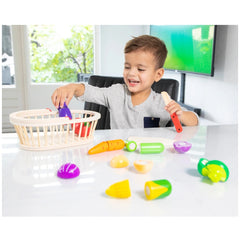 Young child in gray t-shirt seated at a white table with a wicker basket containing red and purple objects, holding green and