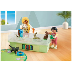 Playmobil Rabbit Family Playset features two small figurines on a clear tabletop, surrounded by colorful books and a blue bow