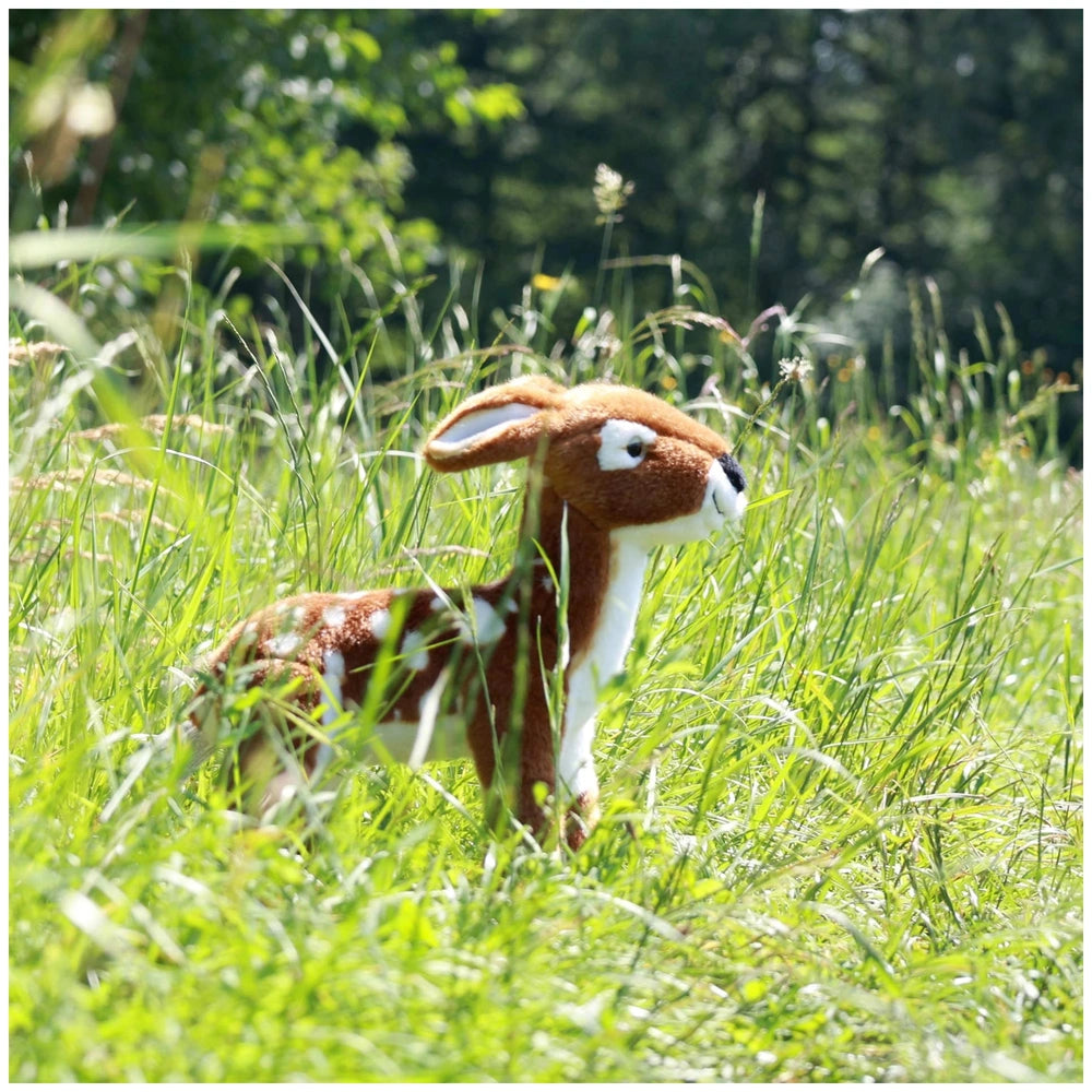 Plush fawn toy standing in a lush green field, showcasing its brown and white color scheme with alert ears and expressive fea