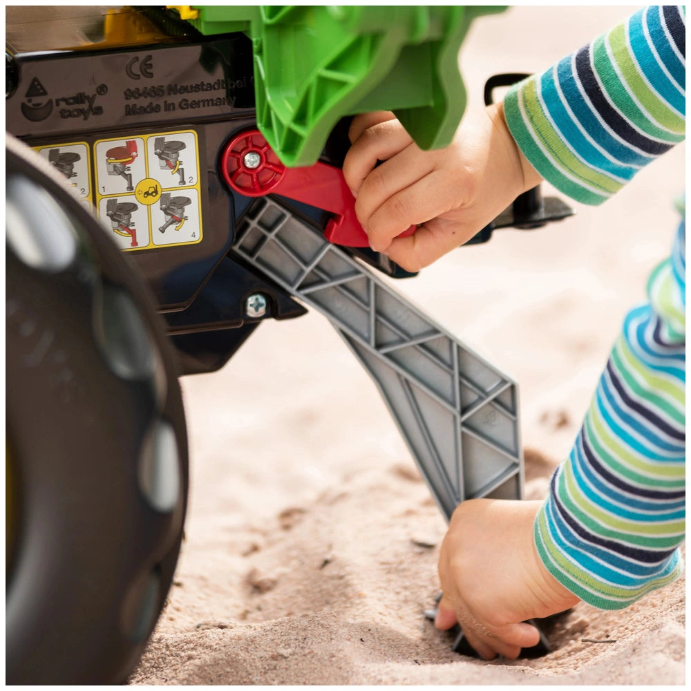 Rollybackhoe John Deere toy features a child's hands pushing a red lever on its metallic structure, partially covered in gree