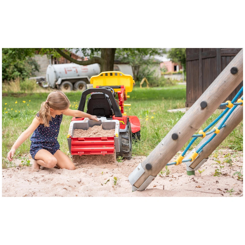 Rollybox John Deere in red and gray sits in the sand as a young girl plays nearby, surrounded by green plants and a wooden st