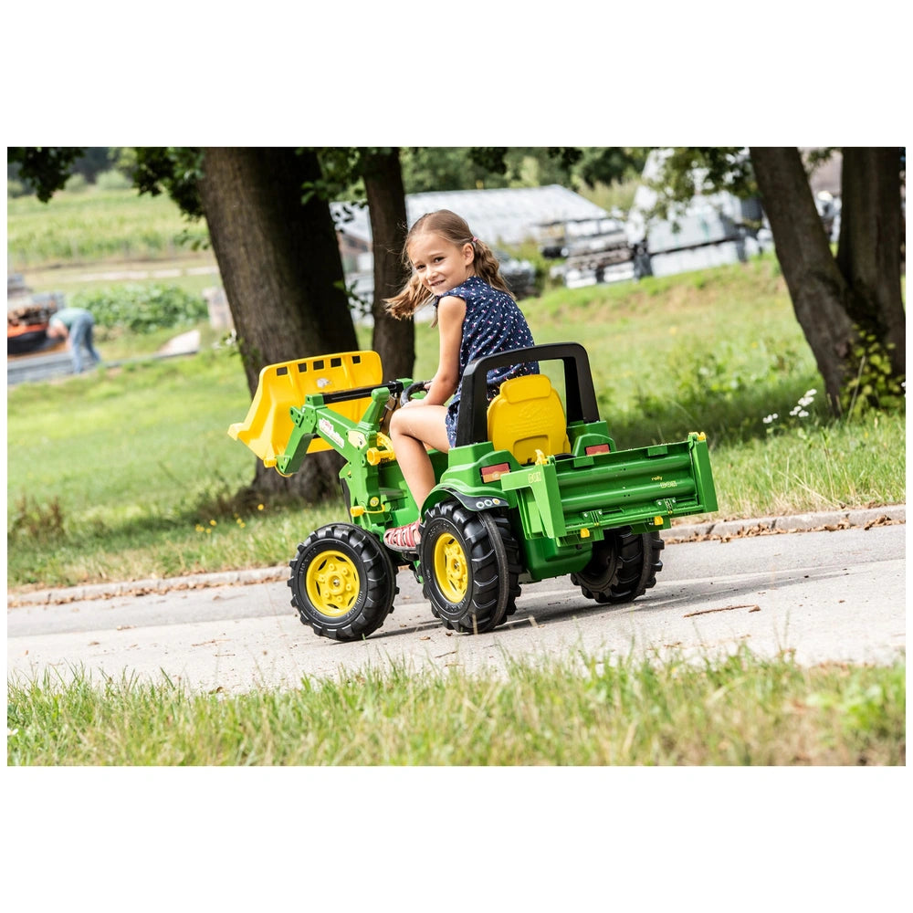 Green and yellow Rollybox tractor with a young girl riding, set on a paved road surrounded by grass and trees.