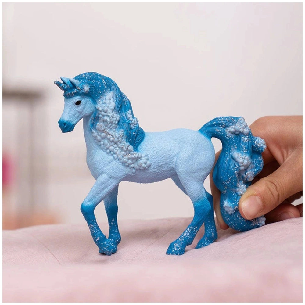 Blue toy water unicorn with white speckles, held by a hand against a soft pink fabric background.