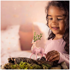 Pink toy deer with green antlers held by a young girl against a light-colored textured pillow in a lifestyle scene.