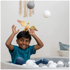 A child in a blue t-shirt smiles while sitting in a white container filled with balls, wearing a toy bird on their head.