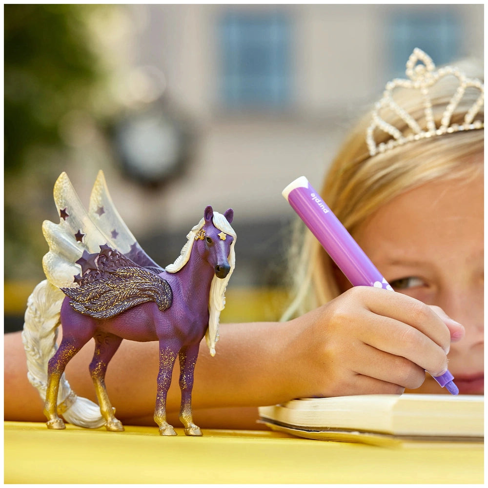 Blonde girl in a white dress holds a purple and gold Schleich Star Pegasus Mare toy beside a yellow table and a book.