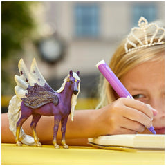 Blonde girl in a white dress holds a purple and gold Schleich Star Pegasus Mare toy beside a yellow table and a book.