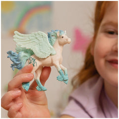 Schleich Stormy Unicorn Foal figurine with glossy white body, textured blue mane, wings, and hooves held in a hand.