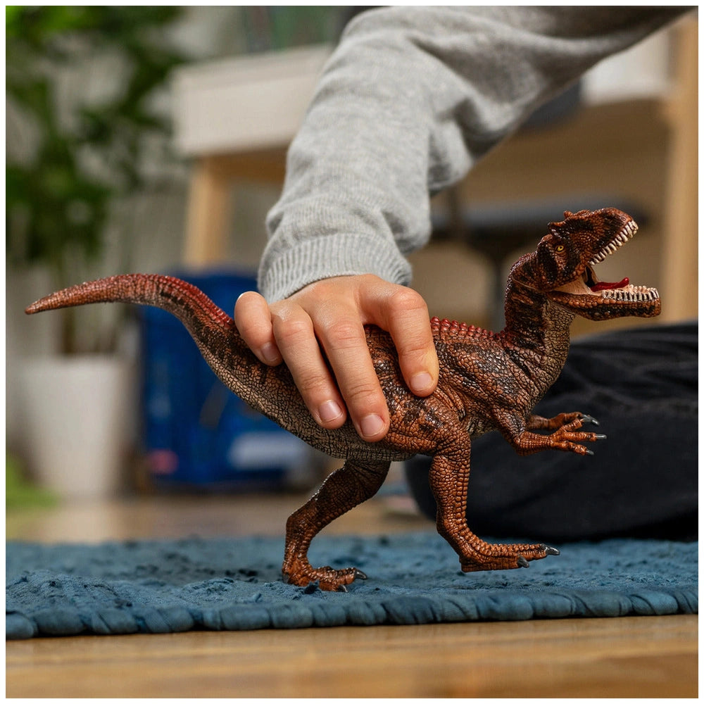 Allosaurus toy dinosaur with textured scaly skin held by a person's hand on blue fabric background.