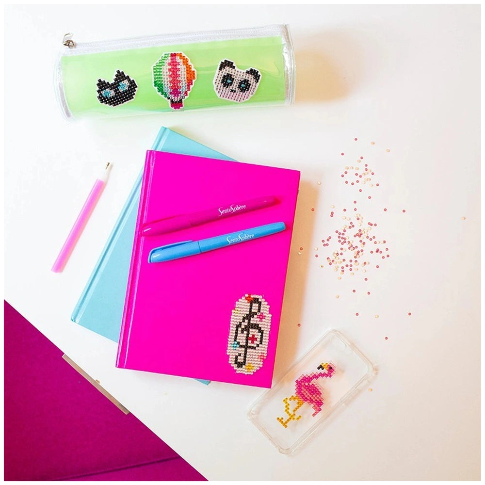 Diamond stickers are surrounded by a pink notebook, blue pen, pencil, and a clear container on a white surface.