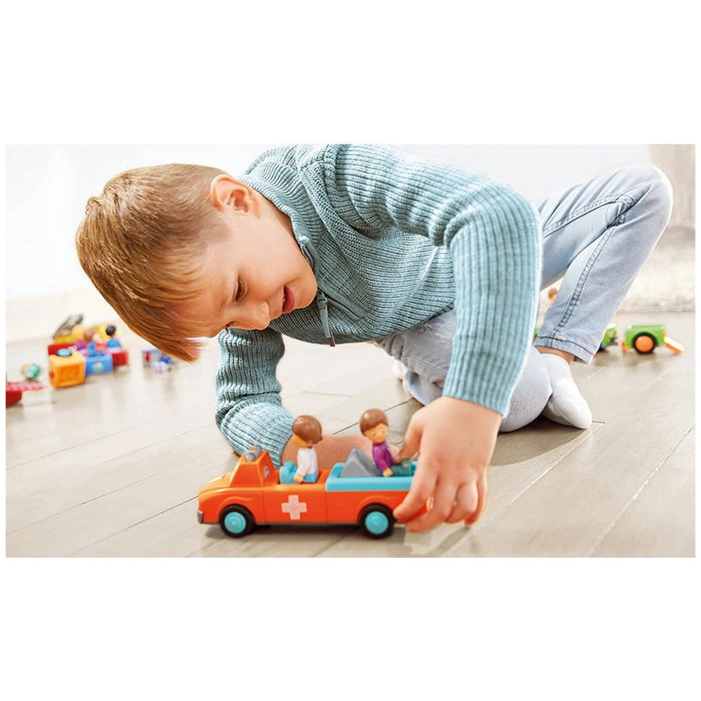 A young boy in a blue sweater and jeans plays with a red and blue toy ambulance car on a wooden floor.