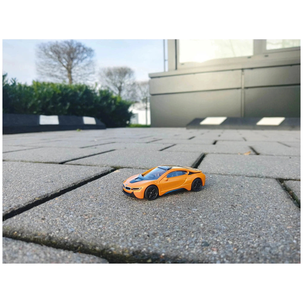 Orange BMW I8 toy car on a paved surface with green shrubs and a reflective building in the background under an overcast sky.