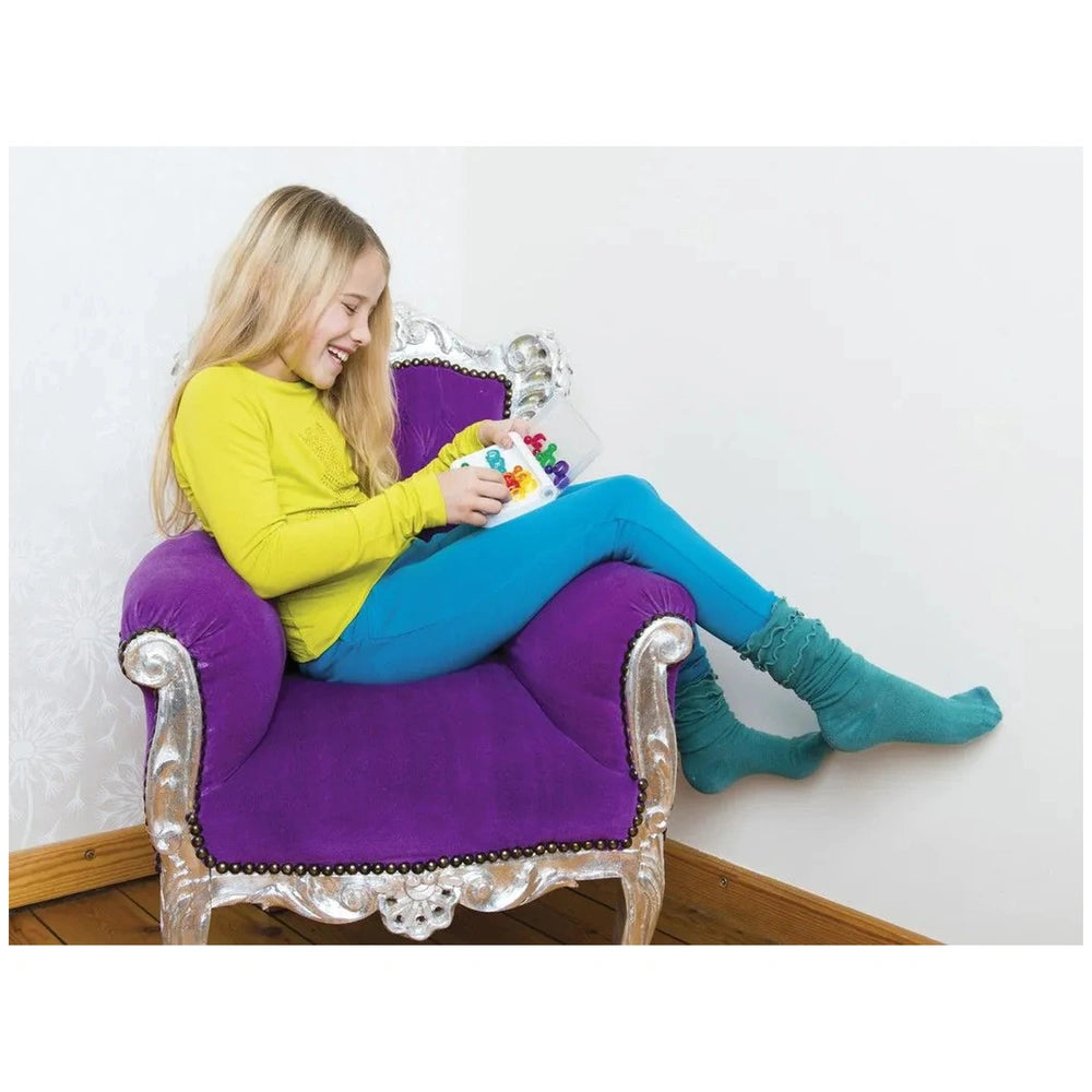 Smart Games IQ Link puzzle is held by a smiling girl in a yellow shirt and blue leggings, seated on a purple velvet chair.