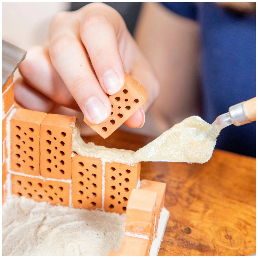 Teifoc beach house construction project features hands applying adhesive to an orange brick on a wooden surface.