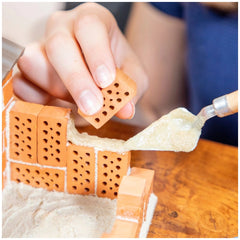 Teifoc beach house construction project features hands applying adhesive to an orange brick on a wooden surface.
