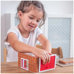 A young girl in a white top smiles while holding a small red and white toy fire station at a wooden table.