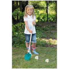Young girl in a white shirt and blue rain boots holds a green and white shovel in a grassy area with flowers and a wooden fen