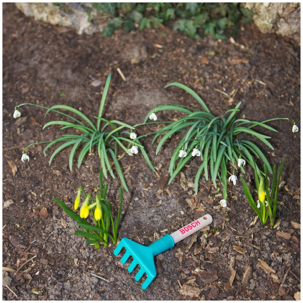 Theo Klein Bosch Garten hand rake in green with metal tines, surrounded by yellow and white plastic flower petals on dirt.
