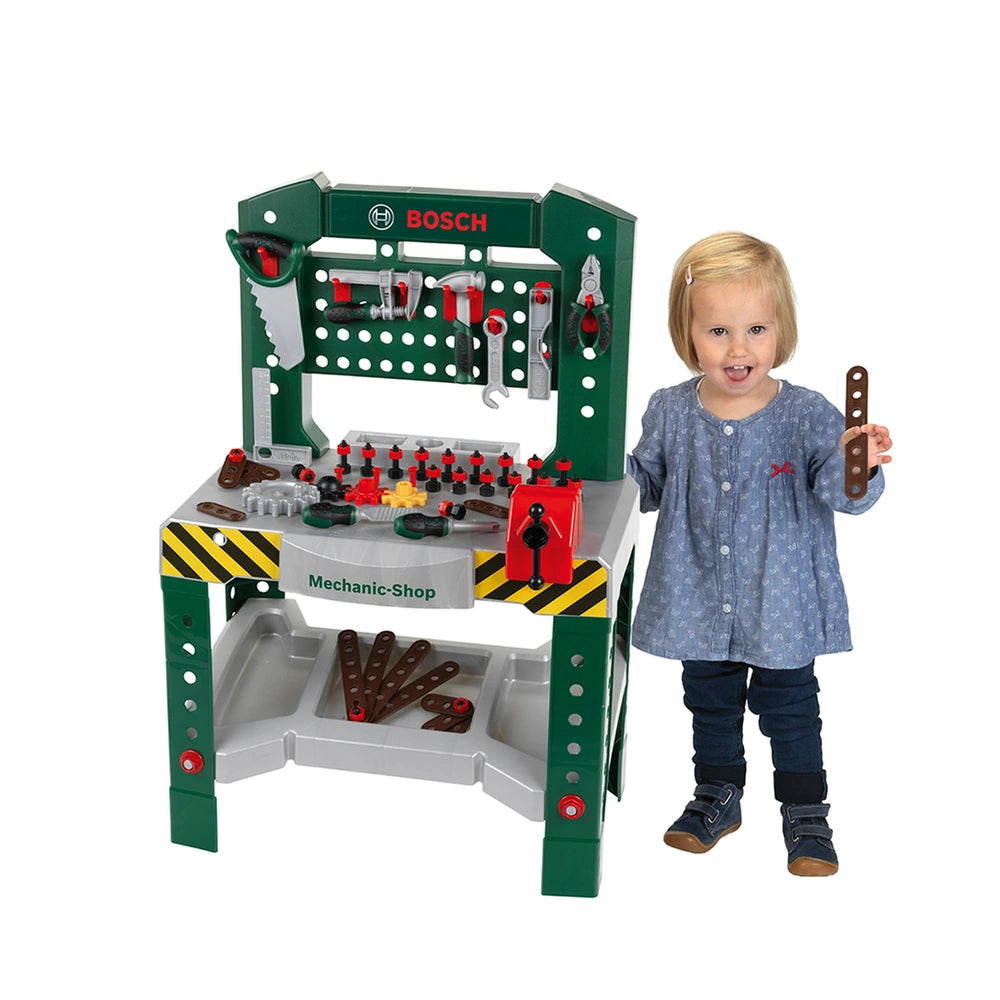 Theo Klein Bosch workbench with green base, white top, and child in blue dress holding a brown tool, surrounded by toy tools.